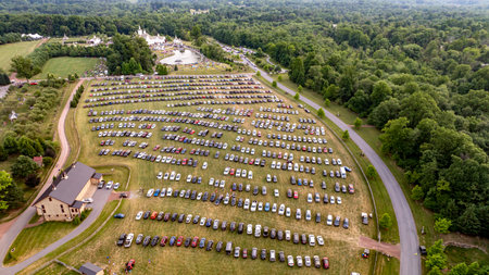 Expansive Parking Area Filled With Cars Leading To A Rural Fairground With White Barns, Small Churches, And A Scenic Pond, Surrounded By Lush Greenery And Rolling Hills.の写真素材