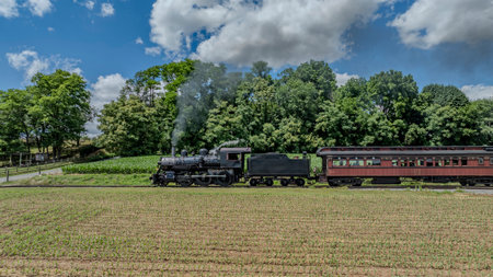 A Black Steam Locomotive Pulling Red Passenger Cars Passes Through A Verdant Countryside With Lush Green Trees In The Background, Billowing Smoke Against A Partly Cloudy Sky.の写真素材