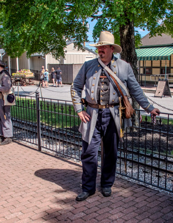 Strasburg, Pennsylvania, USA, June 8, 2024 - A Civil War Reenactor In A Gray Military Coat With Gold Accents And A Beige Hat Stands Casually By A Black Iron Fence Near Railroad Tracks, With Trees And Modern Buildings In The Background Under A Clear Sky.のeditorial素材