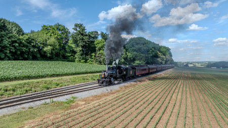 Ronks, Pennsylvania, USA, June 8, 2024 - A Black Steam Locomotive Billowing Thick Smoke Pulls Red Passenger Cars Through Lush Green Fields And Forested Areas, Under A Clear Blue Sky With Puffy Clouds, Creating A Picturesque Countryside Journey.のeditorial素材