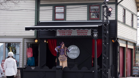 Strasburg, Pennsylvania, USA, April 5, 2024 - A Performer Dressed In A Purple And Gold Costume Stands On An Outdoor Stage Labeled Town Square Stage, Engaging With The Audience. The Stage Is Decorated With Red Curtains And A Sign Listing Show Times, Set Agのeditorial素材