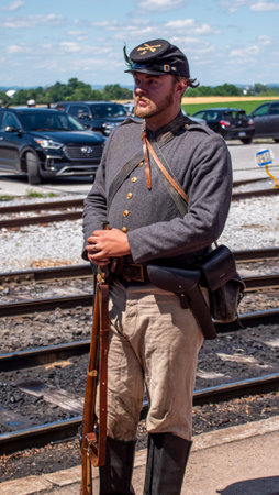 Strasburg, Pennsylvania, USA, June 8, 2024 - A Civil War Reenactor In A Gray Military Uniform And Black Cap Stands By Railroad Tracks, Holding A Rifle And Displaying A Serious Expression, With Modern Cars And A Clear Blue Sky In The Background.のeditorial素材