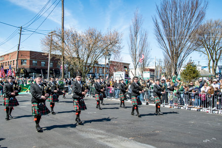 Lindenhurst, New York, USA, March 30, 2024 - Group Of Bagpipers Wearing Traditional Scottish Kilts And Black Jackets March Down A Sunlit Street During A Parade, With A Large Crowd Of Onlookers And Trees In The Background, Celebrating A Cultural Festival.のeditorial素材