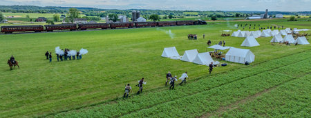 Ronks, Pennsylvania, USA, June 8, 2024 - Civil War Reenactors Perform A Battle Scene On A Green Field Next To Rows Of White Tents, With A Vintage Train In The Background And Rolling Hills Under A Cloudy Sky.のeditorial素材