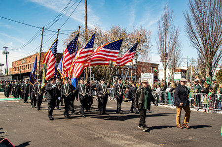 Lindenhurst, New York, USA, March 30, 2024 - Uniformed Marchers Carrying American Flags Walk Proudly Down A Sunny Street During A Parade, With A Lively Crowd Of Spectators Lined Up On The Sides, Celebrating A Patriotic Event In An Urban Setting.のeditorial素材