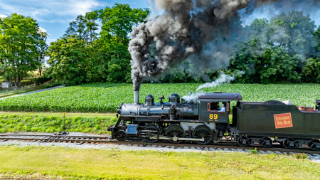 Ronks, Pennsylvania, USA, June 8, 2024 - The Black Steam Locomotive Number 89, Emitting Thick Black Smoke, Travels On Railroad Tracks Beside Verdant Green Crops And Dense Forest, Under A Clear Blue Sky With Puffy Clouds, Showcasing A Picturesque Scene.のeditorial素材