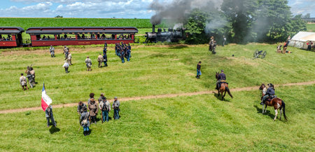 Ronks, Pennsylvania, USA, June 8, 2024 - People In Civil War Uniforms Stand On A Grassy Field Next To A Steaming Locomotive, Engaged In A Reenactment, With Horses, Tents, A Cannon, And A Historical Flag Under Clear Blue Skies.のeditorial素材