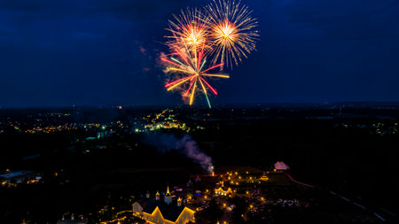Brilliant Gold And Colorful Fireworks Burst Against The Night Sky, Illuminating A Rural Fairground With White Barns And Small Churches, Enhancing The Festive Atmosphere Amidst The Dark Landscape.の写真素材