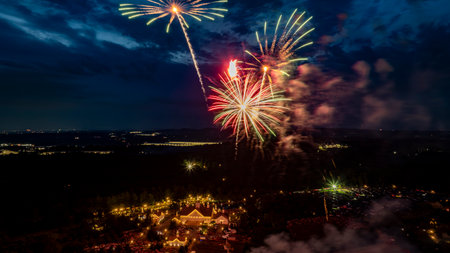 Bright Green And Red Fireworks Explode Over A Well-Lit Estate Amidst Lush Trees, With City Lights In The Distance, Creating A Mesmerizing Nighttime Celebration Scene Against A Dark Sky.の写真素材