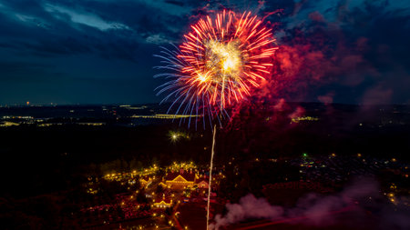 Bright Red And Gold Fireworks Light Up The Night Sky Over A Picturesque Rural Fairground With White Barns And Small Churches, Creating A Festive And Vibrant Atmosphere Against The Dark Backdrop.の写真素材
