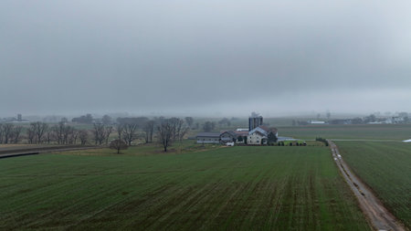 A rural farmstead is enveloped in fog, surrounded by expansive green fields and distant structures on a calm morning.の写真素材