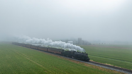 A steam locomotive pulls several carriages along a misty track, passing through lush green fields,...