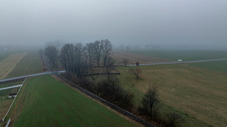 A misty morning covers a rural area, highlighting scattered trees and expansive fields, with a road partially visible.の写真素材