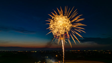 Golden Fireworks Exploding In The Night Sky, Forming A Large, Radiant Burst Over The Distant City Lights And A Gathered Crowd, With The Twilight Horizon Enhancing The Festive Atmosphere.の写真素材