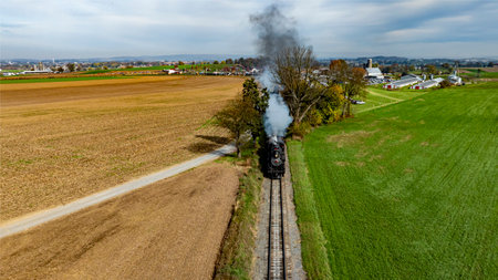 A steam train moves along the tracks beside fields and farms, with smoke billowing into the clear sky on an autumn afternoon.の写真素材