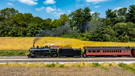 A steam train chugs along the railway, passing vibrant golden fields under a clear blue sky.の写真素材