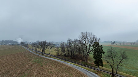A foggy morning reveals a winding railway bordered by barren trees in a tranquil rural countryside. The landscape shows muted colors under overcast skies, creating a serene atmosphere.の写真素材