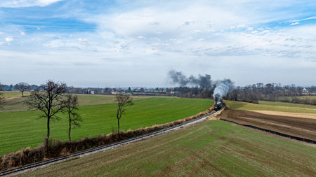 A steam locomotive chugs along a winding track surrounded by lush green fields and distant trees....