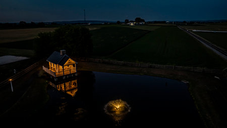 Tranquil Farm Scene At Dusk Featuring A Cozy Stone House Illuminated By Soft Lighting, Reflecting In A Peaceful Pond With A Fountain, Surrounded By Expansive Green Fieldsの写真素材