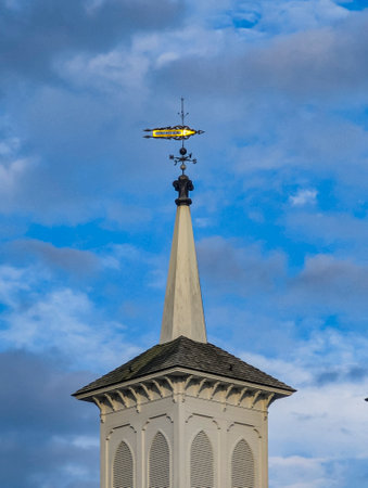 A yellow blimp hovers gracefully above a tall white church steeple, with a backdrop of blue sky and scattered clouds, capturing a serene afternoon atmosphere.の写真素材