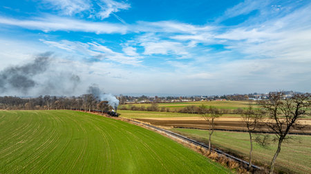 A train emits smoke as it travels through lush green fields. The landscape is expansive, with distant trees and a small town visible under a blue sky marked with soft clouds.の写真素材