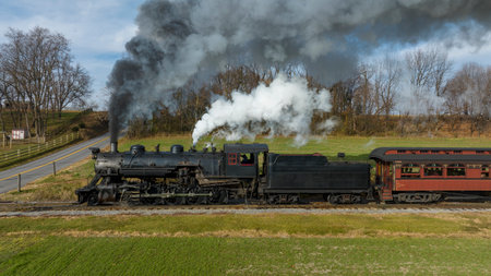 A classic steam locomotive is moving along tracks surrounded by lush green fields and trees, billowing smoke into the clear blue sky on a beautiful day.の写真素材