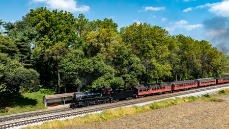 A vintage steam locomotive pulls a charming passenger train along the tracks, surrounded by vibrant trees and a clear blue sky. It creates a nostalgic atmosphere on this beautiful day.の写真素材