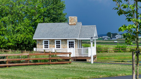 A quaint stone cottage stands in a lush green field, with a clear blue sky and a few clouds. The spacious property features a wooden fence and a scenic landscape beyond.の写真素材