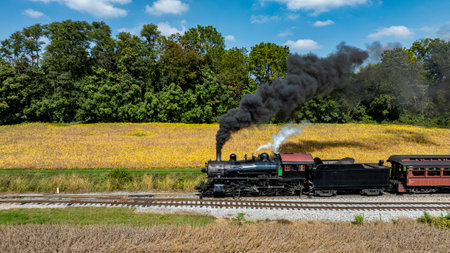 A steam locomotive emits black smoke as it moves along the tracks near a golden field and trees under a clear blue sky.の写真素材