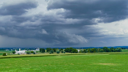 Dark clouds loom ominously over a vast green field, hinting at an approaching storm. A farm can be seen in the distance, framed by the lush countryside as day turns to dusk.の写真素材
