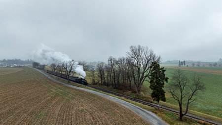 A vintage steam train glides along railway tracks bordered by fields and trees under a cloudy,...