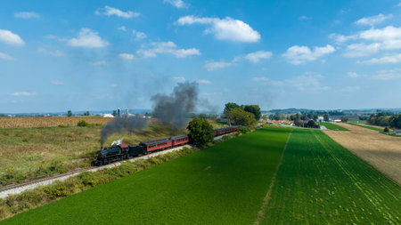 A classic steam locomotive pulls vintage carriages as it travels through vibrant green fields under a bright blue sky, providing a glimpse of nostalgic train journeys in the countryside.の写真素材