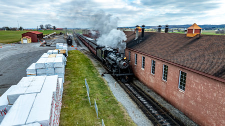 A historic steam locomotive puffs clouds of smoke as it moves next to a brick building. Nearby,...