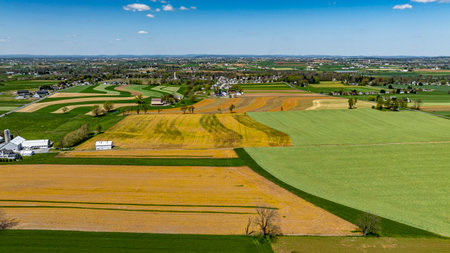 A stunning view of rolling farmland reveals fields of varying colors, agricultural patterns, and a distant rural community, all bathed in sunlight under a bright blue sky.の写真素材