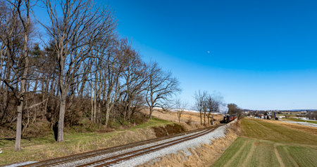 A vintage steam locomotive is making its way along the railway, surrounded by trees and rolling hills. The sky is clear and offers a stunning view of the scenery.の写真素材