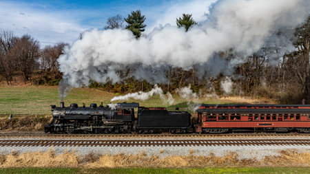 A classic steam locomotive billows smoke as it travels along the railway, framed by greenery and clear skies, showcasing the beauty of a bygone era in transportation.の写真素材