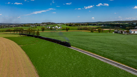 A vintage steam train winds its way through expansive green fields, with gentle hills in the...