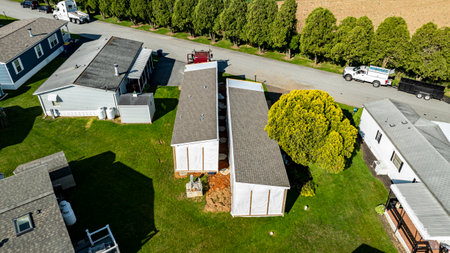 Aerial view shows mobile, manufactured homes lined along a tranquil street, surrounded by well-maintained lawns and trees. The sun casts a warm light, highlighting the neat layout and construction. . High quality photoの写真素材