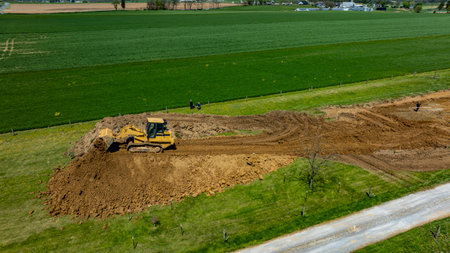 Construction equipment is actively moving earth on a rural property surrounded by lush green fields, with workers laboring nearby under a clear blue sky.の写真素材