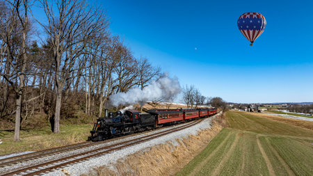 A vintage steam locomotive chugs along the tracks, releasing white smoke as it journeys through lush greeneryの写真素材