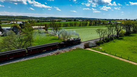 A steam locomotive moves along tracks bordered by vibrant green fields and trees under a clear blue sky. The scenic view captures the charm of countryside rail travel.の写真素材