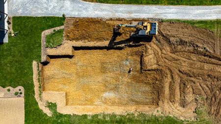 An excavator is digging into the ground to prepare a foundation for building a new structure in a residential neighborhood. The area is clear, showcasing the construction site layout.の写真素材