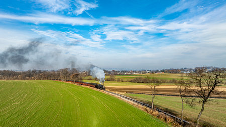 A vintage steam locomotive chugs along tracks bordered by vibrant green fields, surrounded by distant hills and trees under a bright, sunny sky.の写真素材