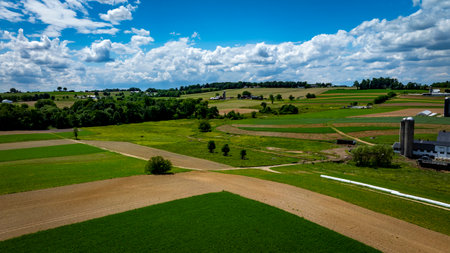 Green fields and brown patches extend across the countryside, framed by trees and a distant hilly backdrop. Bright clouds float above, casting shadows on the vibrant landscape during daytime.の写真素材