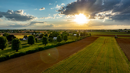 As the sun sets in a vibrant sky, vast agricultural fields stretch across the landscape, dotted with trees and farm structures, creating a serene and picturesque rural scene.の写真素材