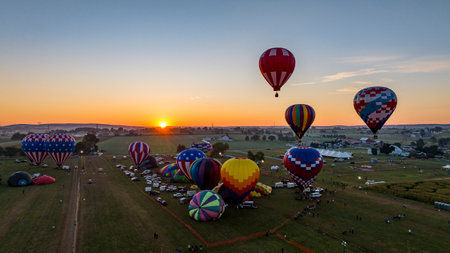 A dozen colorful hot air balloons are preparing for launch in a festival field during sunset. Spectators gather to witness the spectacle as the evening sky radiates warm colors.の写真素材