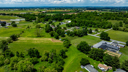 Rolling green fields stretch across the landscape, dotted with trees and farmland. A distant barn and homes are visible, showcasing serene rural life and agricultural beauty.の写真素材
