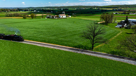 A vintage steam train chugs along a winding track, surrounded by vibrant green fields and rustic farmhouses. The sun shines brightly, highlighting the picturesque rural landscape.の写真素材