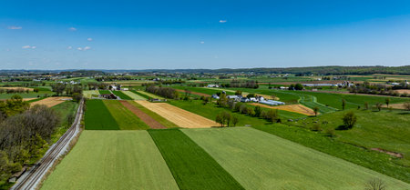 A sweeping view reveals expansive patchwork fields in vibrant green and gold, set against a clear blue sky. A railway line runs along one edge, highlighting the rural charm of the area.の写真素材