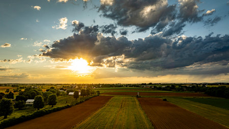 A vibrant sunset casts warm colors over expansive farmland, with lush green fields and patches of clouds creating a picturesque sky. The serene landscape reflects the tranquility of rural life.の写真素材